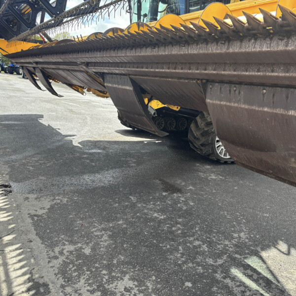 Close-up side view of a large yellow farming harvester header with sharp teeth, parked on asphalt with tractors in the background