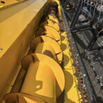 Close-up view of a yellow agricultural machine showing a long yellow auger screw inside its housing, with metal side panels and a row of black-framed tines on the right edge.