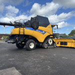 Side view of a large yellow New Holland CR8.90 combine harvester with a long black header parked in a lot under a blue sky.