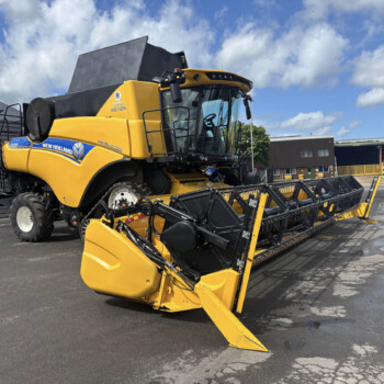 Yellow New Holland combine harvester parked in a yard under a blue sky with clouds.