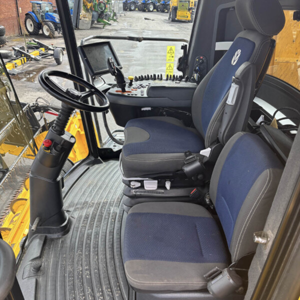 Inside the cab of a construction vehicle, showing the driver's seat, steering wheel, and control panel with levers and buttons, plus the windshield overlooking a yard of tractors and machinery.