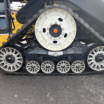 Close-up side view of a heavy tracked machine’s large white drive wheel and rubber track, with yellow machinery in the background and gray body panels behind.