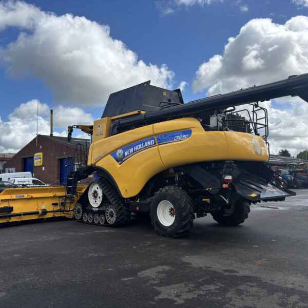 Yellow New Holland CR8.90 combine harvester with its header attached, parked in an equipment yard under a blue sky with clouds.