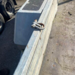 Close-up of a galvanized metal barrier with a rusted bolt and eye screw on top, beside a road and a truck wheel visible in the background.