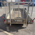 Rear view of a metal-caged utility trailer parked in a lot, with a brick building, a red dumpster, and a white van in the background.