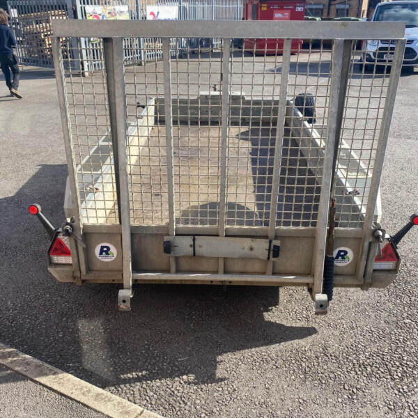 Rear view of a metal-caged utility trailer parked in a lot, with a brick building, a red dumpster, and a white van in the background.
