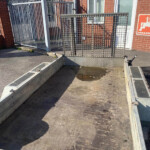 Metal utility trailer with a diamond‑plate floor and raised mesh gate, parked beside a brick building with a white door and windows.