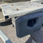 Close-up of a weathered flatbed trailer deck with rusted bolts and a Nugent portable generator mounted at the rear, set against a gravel ground.