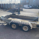Flatbed trailer with hitch and wheels parked in an industrial yard near a gated fence and brick buildings