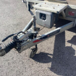 Tow trailer hitch with coiled electrical cables and a gray tool box labeled NUGENT on a sunny asphalt lot