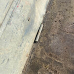 Close-up of a weathered metal panel with bolts, resting against a dirt path; a small cylindrical rod lies at the seam between materials.