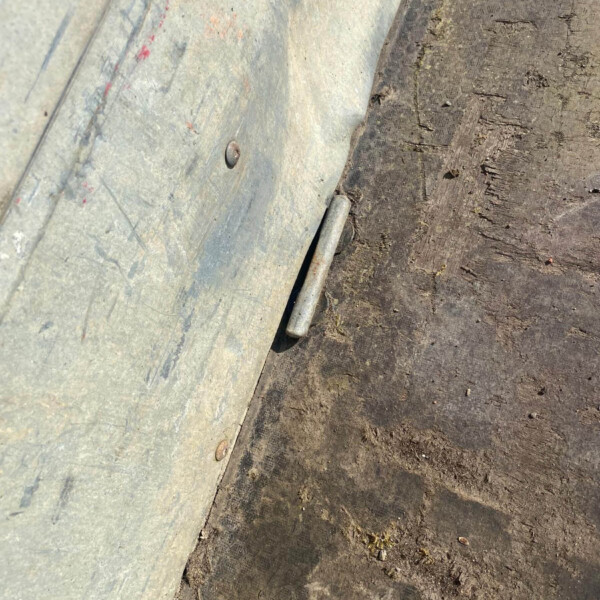 Close-up of a weathered metal panel with bolts, resting against a dirt path; a small cylindrical rod lies at the seam between materials.