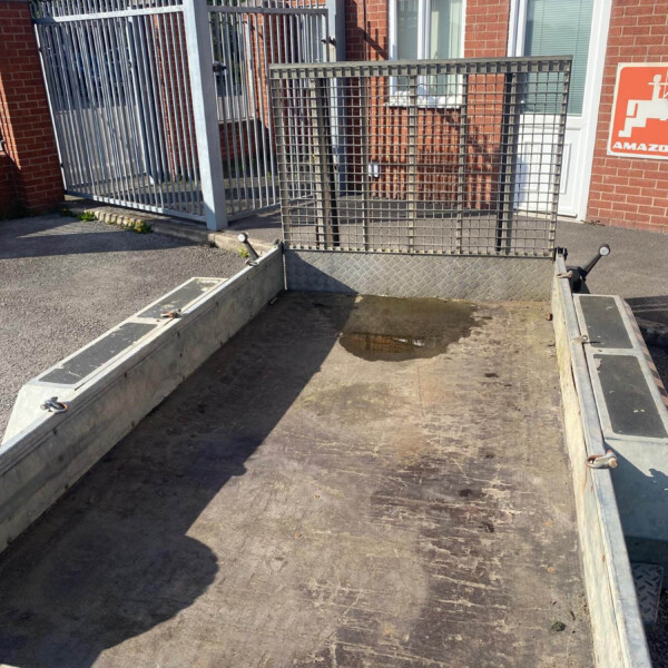 Metal utility trailer with a diamond‑plate floor and raised mesh gate, parked beside a brick building with a white door and windows.