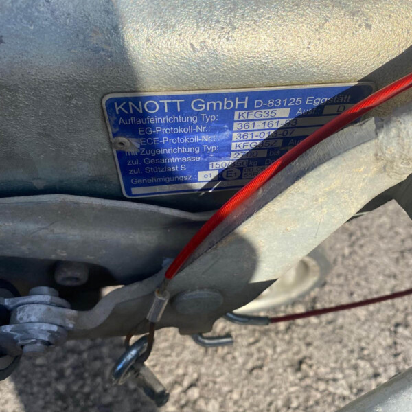 Blue metal identification plate on a trailer frame reading KNOTT GmbH, model and serial numbers, with a red cable crossing in front.