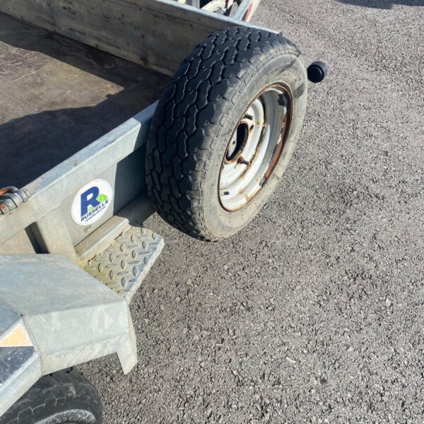 Rear wheel and tire of a metal utility trailer on a gravel surface, showing rust on the rim and a spare mounted nearby.