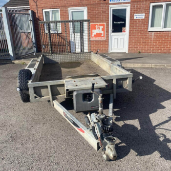 Metal flatbed trailer with a ramp, parked on an asphalt lot in front of a brick building with a reception sign.