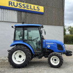 Blue New Holland tractor parked in front of a gray wall with a yellow and blue Russell sign above it.