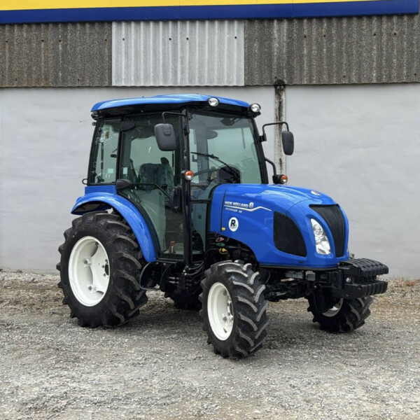 Blue tractor parked on a gravel lot in front of a gray wall with a yellow and blue sign above.