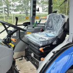 Interior of a tractor cab showing a wrapped seat, steering wheel, and control levers with orange grips; outdoor farm scenery visible through windows