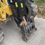 Close-up of a yellow construction loader arm with black fork tines, warning stickers, and a rusted hitch on a concrete surface.