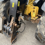 Close-up of a heavy machinery attachment with yellow painted frame, hydraulic hoses, and rusted metal pins near large tire treads, showing warning decals and wear.