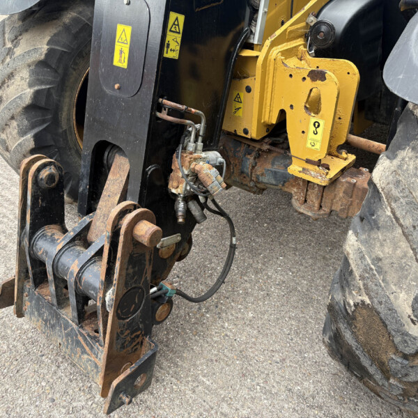 Close-up of a heavy machinery attachment with yellow painted frame, hydraulic hoses, and rusted metal pins near large tire treads, showing warning decals and wear.