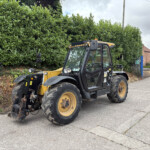 Yellow and black construction vehicle parked along a street beside a tall green hedge, with brick houses in the distance.