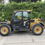 Yellow and black CAT telehandler parked on a paved surface with oversized tires, hedge backdrop behind it.