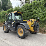 Large CAT telehandler parked on a paved lot beside a tall green hedge, showing its yellow and black body and big tires.