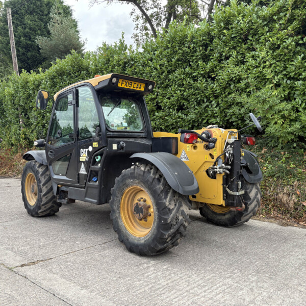 Large CAT telehandler parked on a paved lot beside a tall green hedge, showing its yellow and black body and big tires.