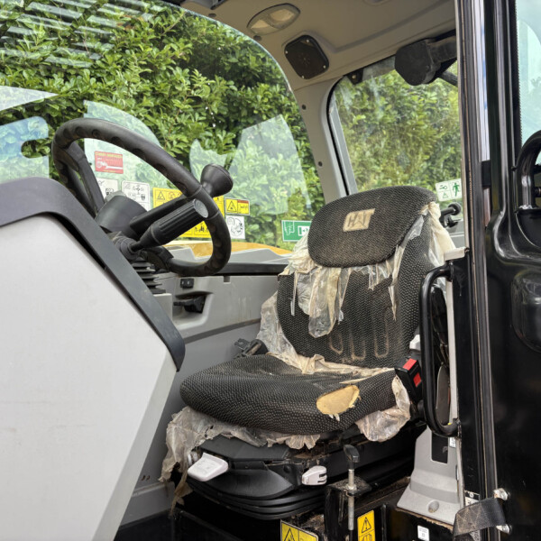 Interior of a construction vehicle cab with a heavily torn fabric seat covered in plastic, showing exposed foam and wear near the base; steering wheel and warning labels visible.