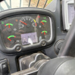 Close-up of a tractor's instrument panel with a central tachometer, green/yellow gauges, and warning lights next to the steering wheel.