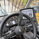 View from the operator seat of a CAT heavy equipment cab showing the steering wheel, dashboard gauges, and a digital display screen with CAT logo and controls nearby.