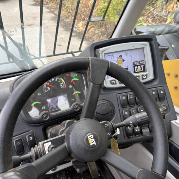 View from the operator seat of a CAT heavy equipment cab showing the steering wheel, dashboard gauges, and a digital display screen with CAT logo and controls nearby.