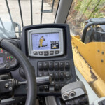 Cab of a heavy construction machine with a CAT control screen showing dig graphics, surrounded by multiple levers and buttons; yellow equipment visible outside the window.