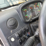 Cockpit of a vehicle showing a round instrument cluster with red and green gauges, steering wheel, and control switches. Dusty dashboard.