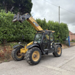 Yellow CAT backhoe/telehandler with its bucket raised over a tall green hedge beside a residential street, with brick houses in the background