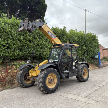 Yellow CAT backhoe/telehandler with its bucket raised over a tall green hedge beside a residential street, with brick houses in the background