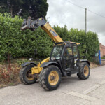 Yellow CAT telehandler with raised bucket reaching over a tall hedge along a residential street on an overcast day.