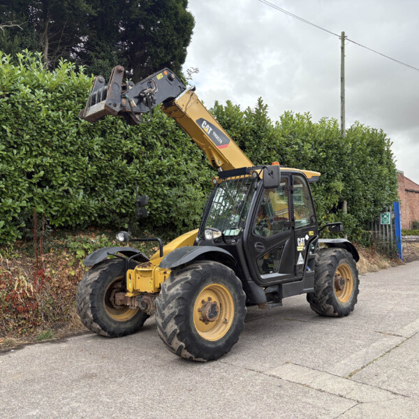 Yellow CAT telehandler with raised bucket reaching over a tall hedge along a residential street on an overcast day.