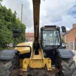 Front view of a yellow excavator parked on a street, with a tall hedge to the left and brick houses in the background.