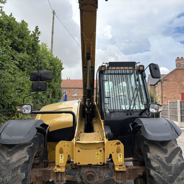 Front view of a yellow excavator parked on a street, with a tall hedge to the left and brick houses in the background.
