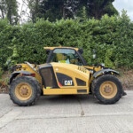 Yellow CAT construction vehicle parked in front of a tall green hedge along a paved area, showing large tires and cabin.