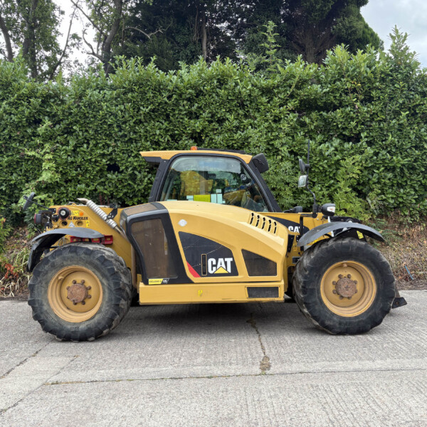 Yellow CAT construction vehicle parked in front of a tall green hedge along a paved area, showing large tires and cabin.