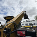 Close-up of a yellow construction excavator arm with a visible license plate FX19 CAA, crane machinery in the foreground and a Russell's building in the background under a cloudy sky.