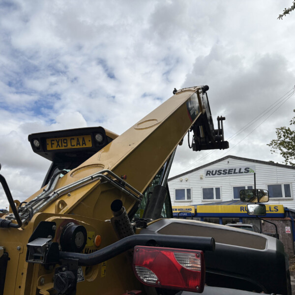 Close-up of a yellow construction excavator arm with a visible license plate FX19 CAA, crane machinery in the foreground and a Russell's building in the background under a cloudy sky.