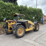 Caterpillar skid-steer loader in yellow parked on a street beside a tall green hedge and brick houses in the background.