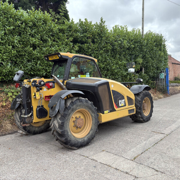 Caterpillar skid-steer loader in yellow parked on a street beside a tall green hedge and brick houses in the background.