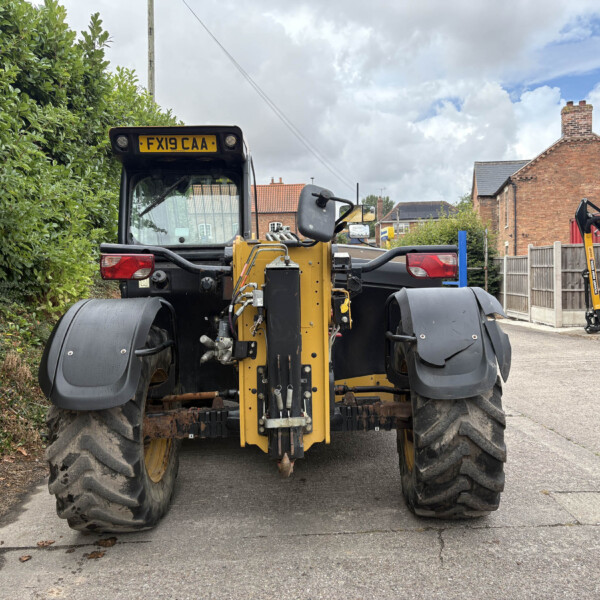 Front view of a large yellow road roller parked on a residential street, with a tall green hedge on the left and brick houses in the background.