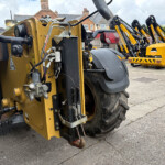 Close-up of a yellow construction machine’s hydraulic components and large wheel, with more yellow machines lined up in the background outdoors.